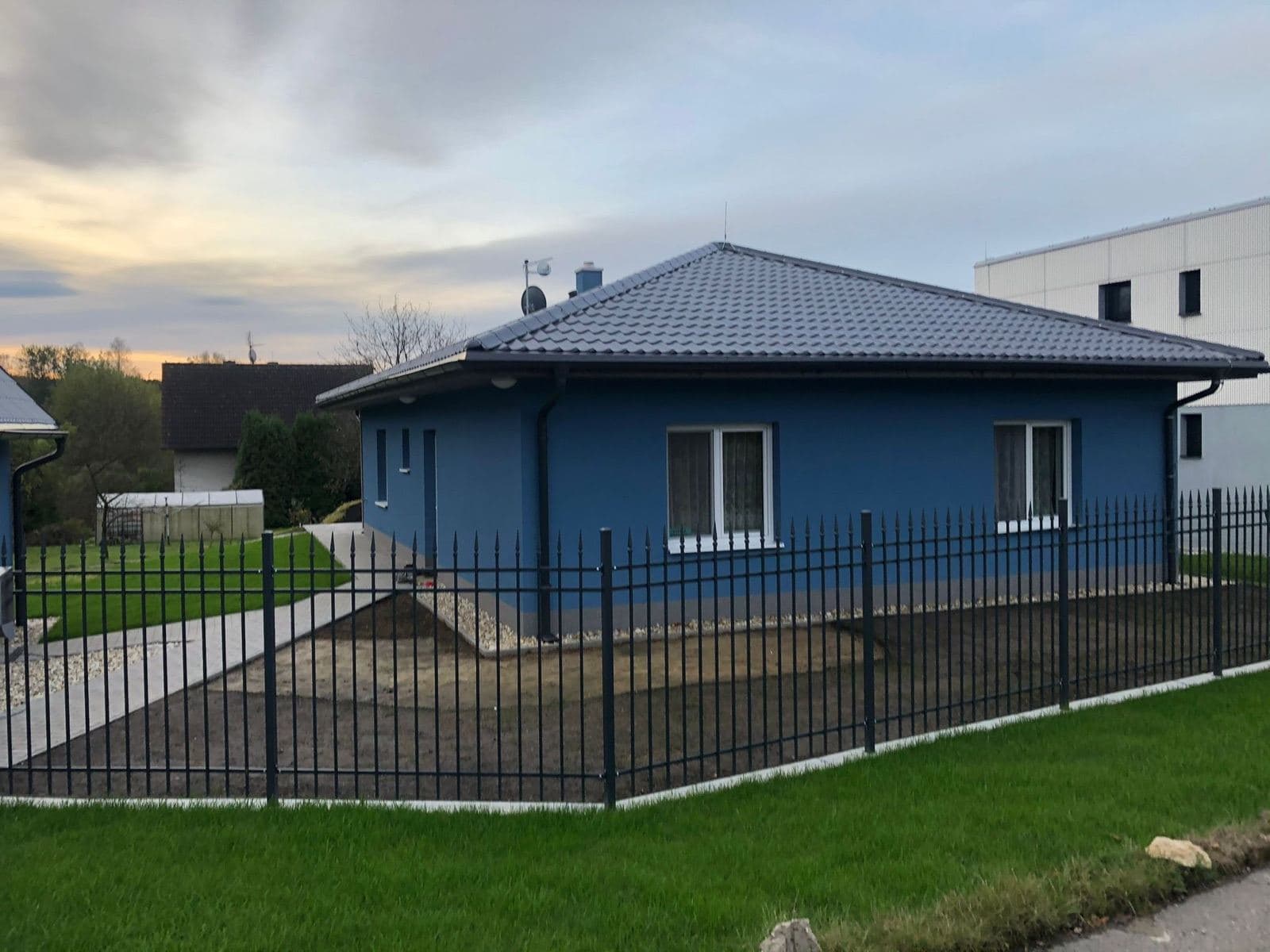 Bungalov s modrou fasádou, valbovou střechou a kovaným plotem při západu slunce