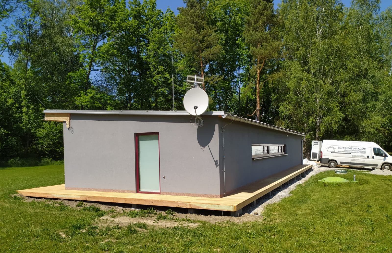 Šedý bungalov s plochou střechou a dřevěnou terasou kolem domu v zahradě
