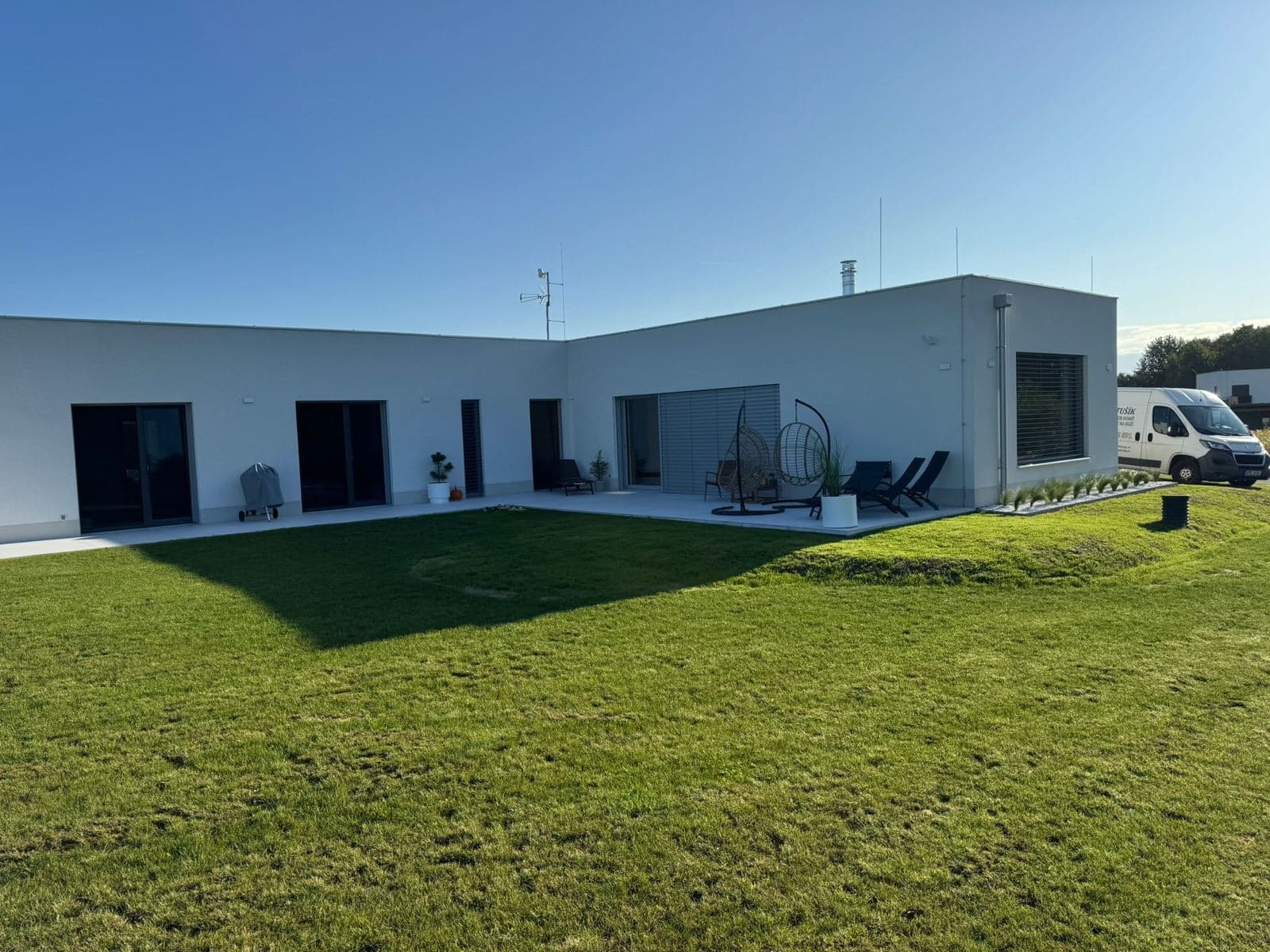 Moderní bungalov s plochou střechou, bílou fasádou a venkovním posezením na dlažbě